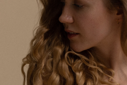 Portrait photograph of a young woman with long, wavy blonde hair. She is looking to the side with a relaxed, downcast gaze.