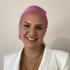 Portrait of a woman with brown skin and short pink hair, wearing a white blazer and smiling brightly.